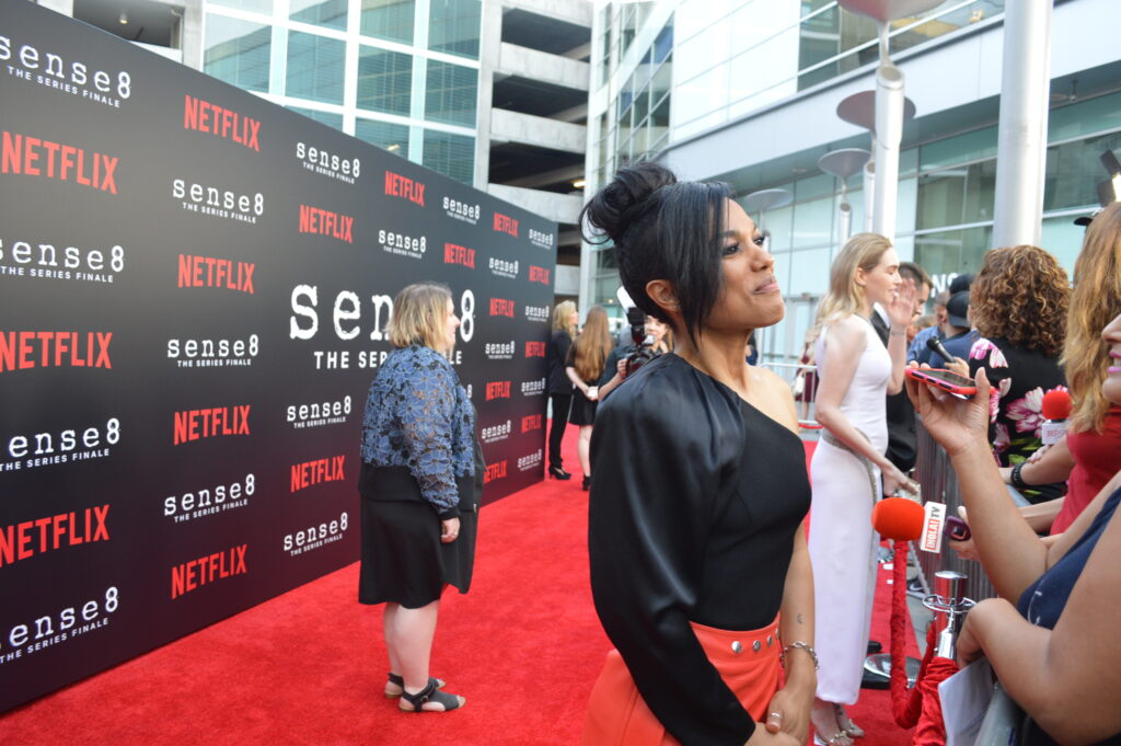 a black woman with black hair, wearing a black top and red skirt, talking to someone with their phone out recording, on the red carpet