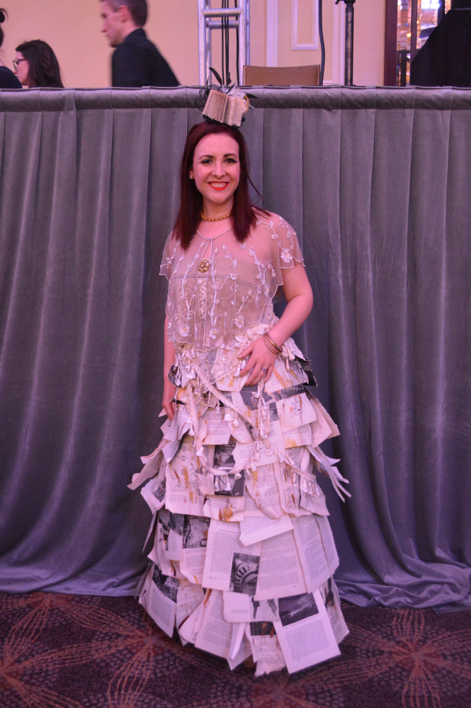 a young white woman with shoulder-length red hair, wearing a prom-style dress made out of book pages