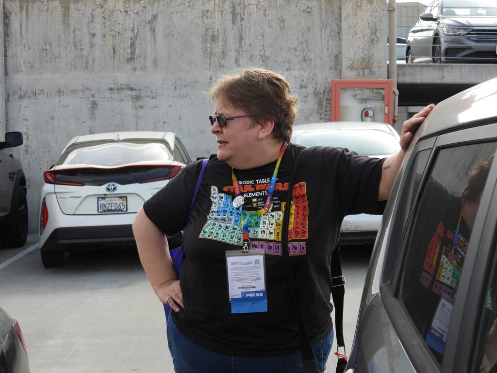 a photo of Angie, wearing a 'periodic table of Star Wars elements' shirt and jeans, standing next to a car, looking away