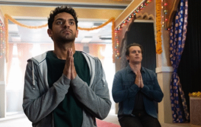 a Hindu temple with yellow walls and a red carpet in the center. In the front left is an Indian man wearing a black top with a gray hoodie kneeling in prayer. Behind him is a white man wearing a blue shirt and black pants, also kneeling in prayer.