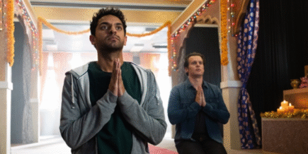 a Hindu temple with yellow walls and a red carpet in the center. In the front left is an Indian man wearing a black top with a gray hoodie kneeling in prayer. Behind him is a white man wearing a blue shirt and black pants, also kneeling in prayer.