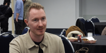 a white man with light brown/reddish hair and a slight mustache wearing a tan button up jacket over a black shirt