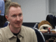 a white man with light brown/reddish hair and a slight mustache wearing a tan button up jacket over a black shirt