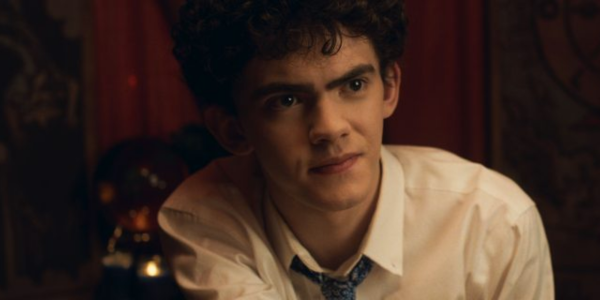 A white teen boy (Joe Locke) with black curly hair and a button up shirt and loose tie, sits looking curious.