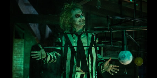 Michael Keaton as Betelgeuse - white and black striped suit, clown white makeup, and wild hair - posing with both hands out