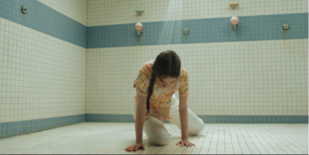 a young white girl with brown hair in a braid, wearing a flowery shirt and white pants, on her hands and knees in a gym shower