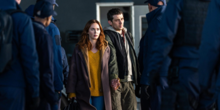 Disclosure Day Review - a white woman wearing a yellow sweater, jeans, and a long brown coat holds the hand of a white man wearing brown pants and a hoodie, surrounded by several men in blue suits