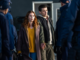 Disclosure Day Review - a white woman wearing a yellow sweater, jeans, and a long brown coat holds the hand of a white man wearing brown pants and a hoodie, surrounded by several men in blue suits