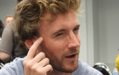 a white man with reddish brown hair and beard, wearing a grey sweatshirt, squinting and pointing to his forehead