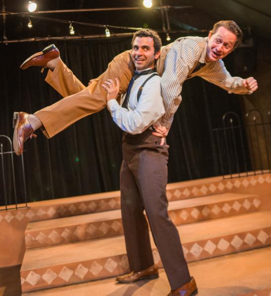 A white man in dark brown slacks and a light blue shirt holds another man in light brown slacks and a tanned shirt over his head. 