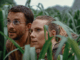 A young white man with brown hair, a slight beard, and glasses crouches next to a young white woman with brown hair back in a ponytail in high grass.