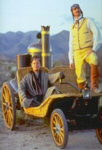a still of John de Lancie and Richard Dean Anderson, wearing western gear, sitting on an old-timey looking vehicle