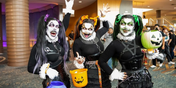 three women, dressed in black outfits and wearing white makeup, pose with trick or treat containers