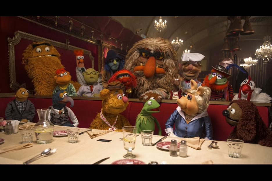 A group of Muppets sit around a restaurant table, some standing behind the seat.