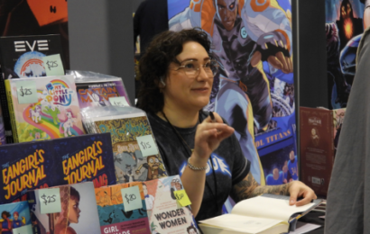 a white woman with short curly brown hair and glasses, sits at a table at a con, ready to get an autograph