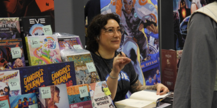 a white woman with short curly brown hair and glasses, sits at a table at a con, ready to get an autograph