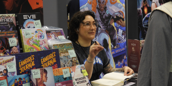 a white woman with short curly brown hair and glasses, sits at a table at a con, ready to get an autograph