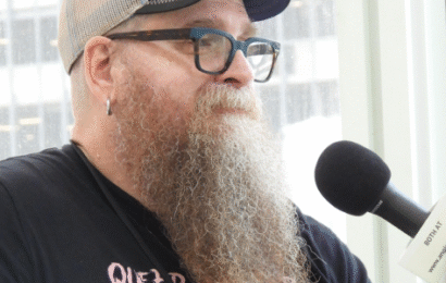 a white man with a long reddish curly beard starting to turn white, wearing glasses, a baseball cap, and a black t-shirt, being interviewed
