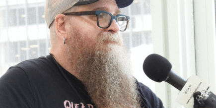a white man with a long reddish curly beard starting to turn white, wearing glasses, a baseball cap, and a black t-shirt, being interviewed