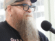 a white man with a long reddish curly beard starting to turn white, wearing glasses, a baseball cap, and a black t-shirt, being interviewed