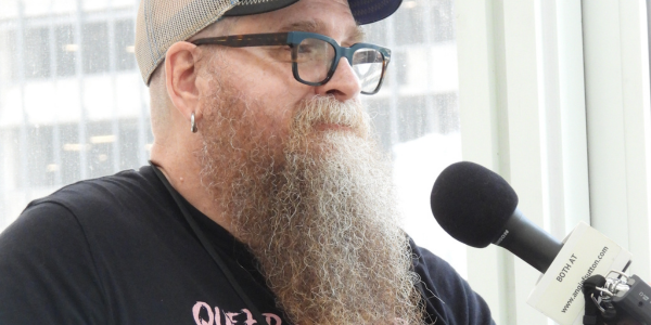 a white man with a long reddish curly beard starting to turn white, wearing glasses, a baseball cap, and a black t-shirt, being interviewed