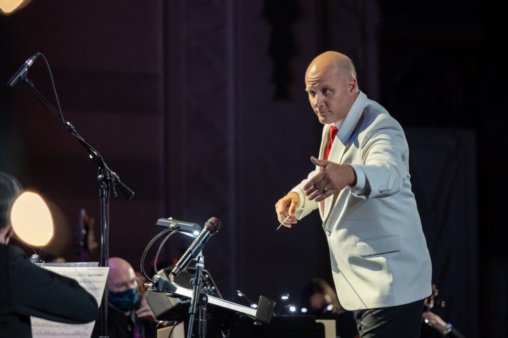 a bald white man in a white jacked conducts an orchestra, you see just the front of the first row