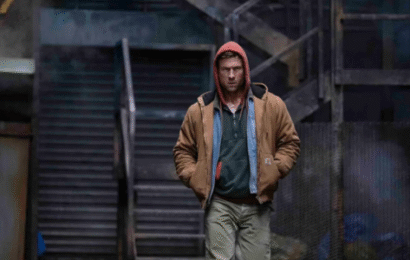 a grungy outdoor location with stairs leading up. A white man wearing a red hood, brown jacket with two shirts underneath, and brown pants is walking toward the screen.