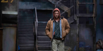 a grungy outdoor location with stairs leading up. A white man wearing a red hood, brown jacket with two shirts underneath, and brown pants is walking toward the screen.