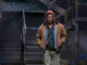 a grungy outdoor location with stairs leading up. A white man wearing a red hood, brown jacket with two shirts underneath, and brown pants is walking toward the screen.