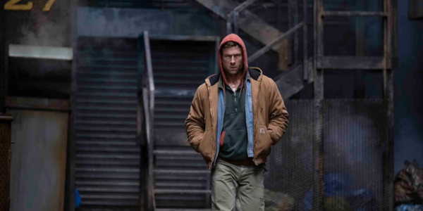 a grungy outdoor location with stairs leading up. A white man wearing a red hood, brown jacket with two shirts underneath, and brown pants is walking toward the screen.