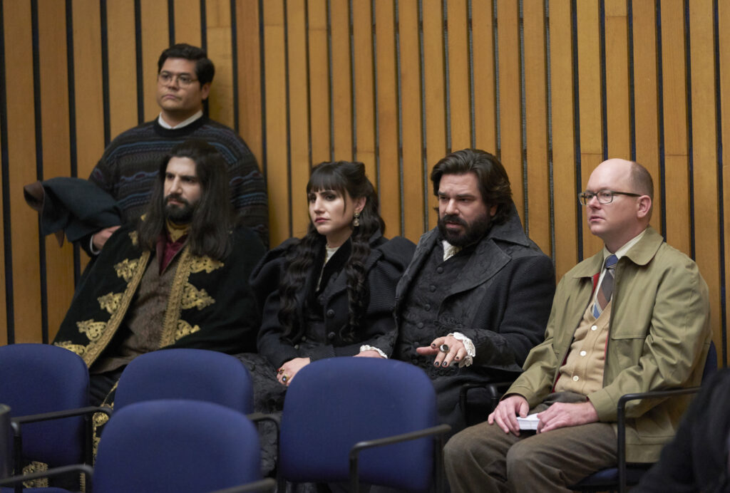 A man standing, and three men and a woman sitting down, all in a room with wooden walls. The standing man is Latinx with black hair and glasses, wearing a sweater and carrying a coat. From left to right, we have a man with long black hair and beard/moustache, wearing a royal blue robe; a woman with long black hair wearing a black fancy dress; a shorter man with short black hair and beard/moustache, wearing a black suit; and a balding man with glasses wearing a tan coat, brown sweater vest, and tie.