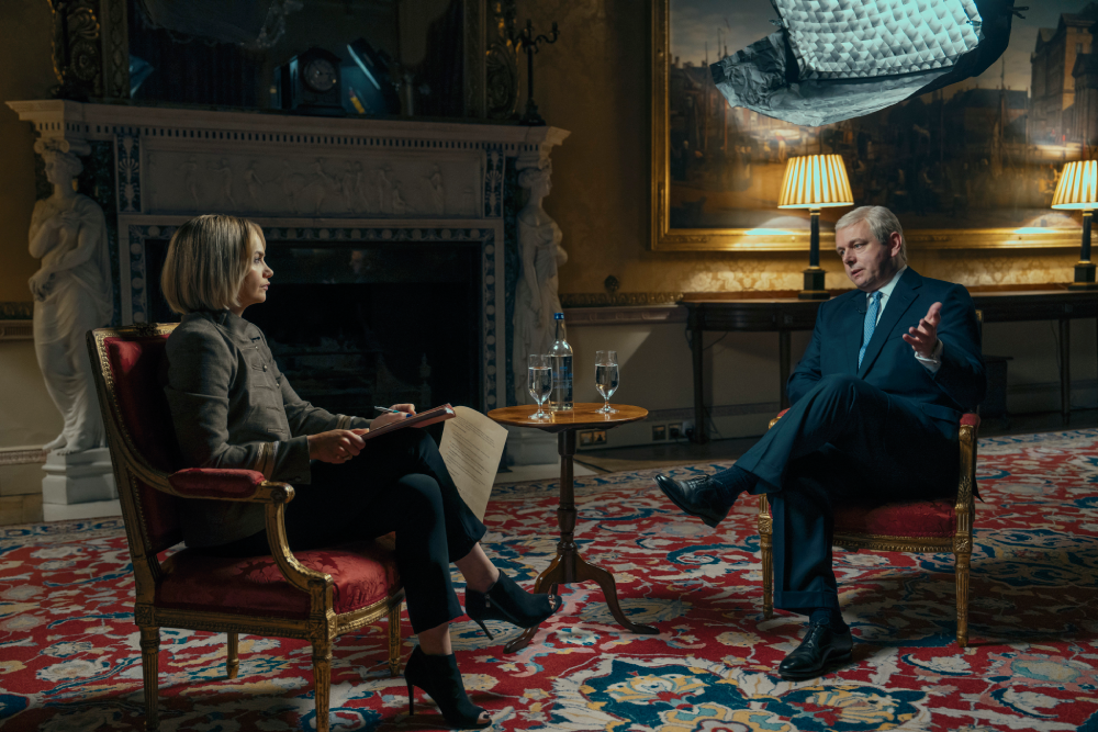 Emily Maitlis (Ruth Wilson: a white woman with blond hair wearing a dark brown coat and pants) and Prince Andrew (Michael Sheen: a white man with white hair wearing a blue suit and tie) sit in two red chairs in an opulent room - obviously the Palace.