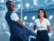 a young black main in a denim kilt sits on a railing while a young black woman wearing jeans and a white shirt stands smiling at him