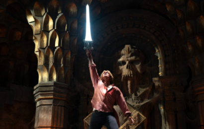 a young white man, wearing a pink shirt and black pants, stands in front of an alter with a skull in it, sword held high