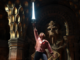 a young white man, wearing a pink shirt and black pants, stands in front of an alter with a skull in it, sword held high