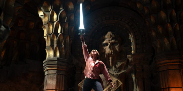 a young white man, wearing a pink shirt and black pants, stands in front of an alter with a skull in it, sword held high